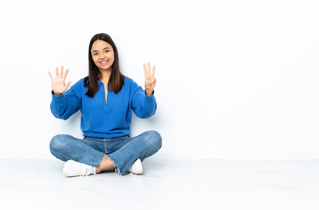 Jeune femme de race mixte assis sur le sol isolé sur mur blanc en comptant huit avec les doigts