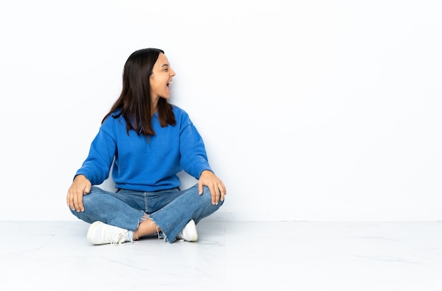 Jeune femme de race mixte assis sur le sol isolé sur mur blanc en riant en position latérale