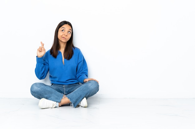 Jeune femme de race mixte assis sur le sol sur le mur blanc avec les doigts se croisant et souhaitant le meilleur