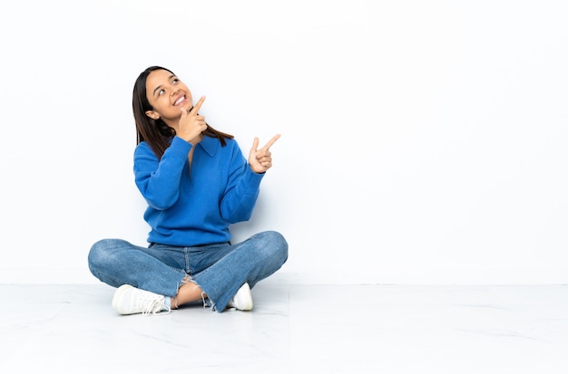 Jeune femme de race mixte assis sur le sol sur le mur blanc pointant avec l'index une excellente idée