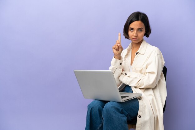 Jeune femme de race mixte assise sur une chaise avec un ordinateur portable isolé sur fond violet comptant un avec une expression sérieuse