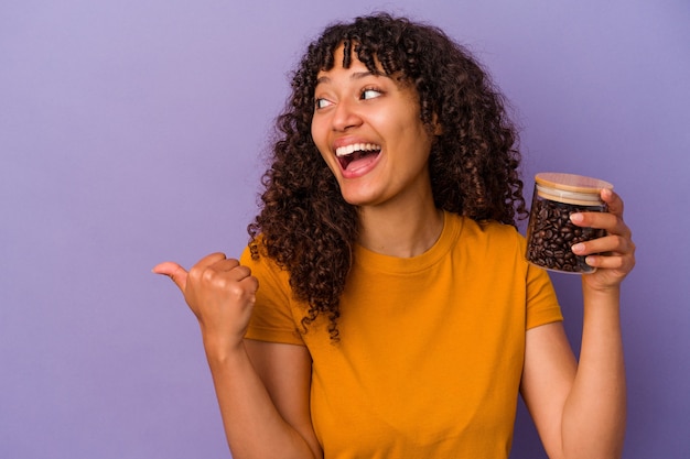 Jeune femme de race mixte tenant une bouteille de grains de café isolée sur des points de fond violet avec le pouce loin, riant et insouciant.