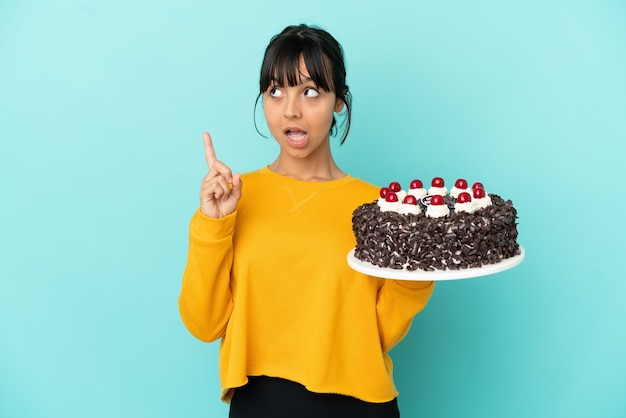 Jeune femme de race mixte tenant un gâteau d'anniversaire pensant à une idée pointant le doigt vers le haut