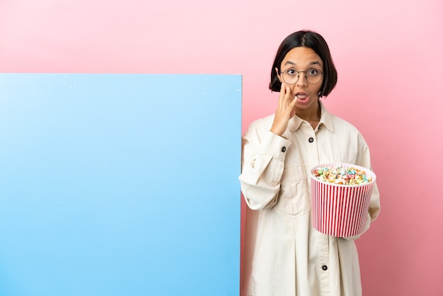 Jeune femme de race mixte tenant des pop-corns avec une grande bannière sur fond isolé avec une expression faciale surprise et choquée