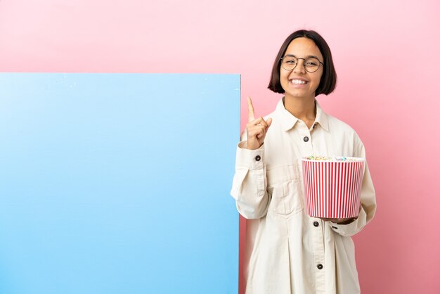 Jeune femme de race mixte tenant des pop-corns avec une grande bannière sur fond isolé pointant vers une excellente idée