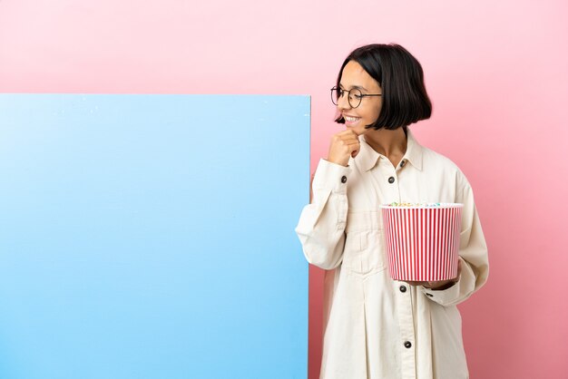 Jeune femme de race mixte tenant des pop-corns avec une grande bannière sur fond isolé regardant sur le côté et souriant