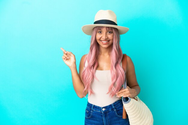 Jeune femme de race mixte tenant un sac de plage avec Pamela isolé sur fond bleu, pointant le doigt sur le côté