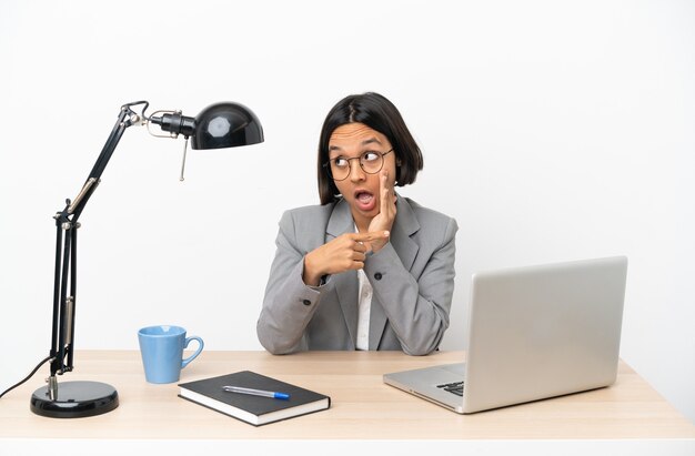 Jeune femme de race mixte travaillant au bureau pointant sur le côté pour présenter un produit et chuchoter quelque chose