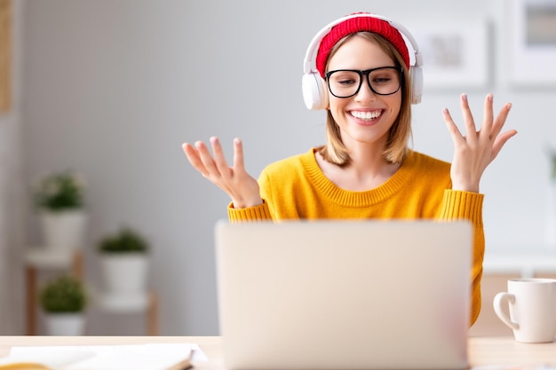 Jeune femme ravie dans des écouteurs et des lunettes souriant et gesticulant assis à table et parlant avec un collègue en ligne à la maison