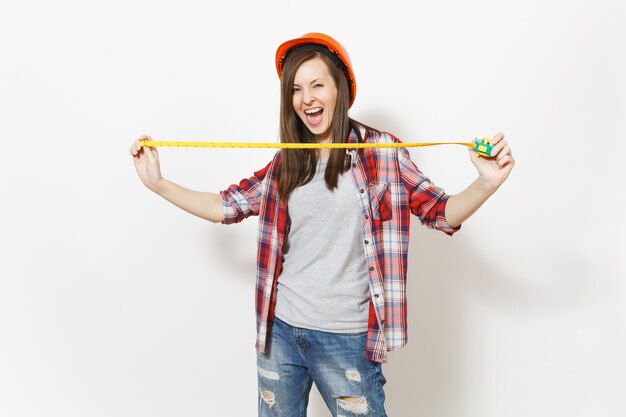 Jeune femme ravie dans des vêtements décontractés, casque de protection de construction tenant un ruban de mesure de jouet isolé sur fond blanc. Instruments, outils pour la salle d'appartement de rénovation. Réparer le concept de maison.