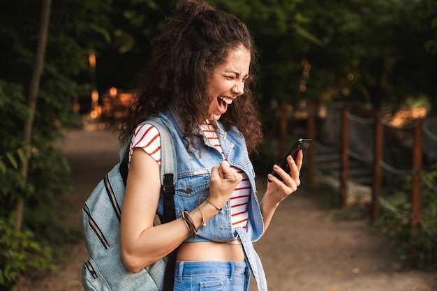 Jeune femme ravie portant sac à dos, serrant le poing et criant avec smartphone à la main tout en marchant dans le parc verdoyant