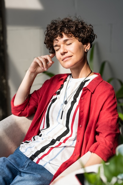 Jeune femme ravie en smart casual assis dans un fauteuil tout en profitant du repos après le travail devant la fenêtre