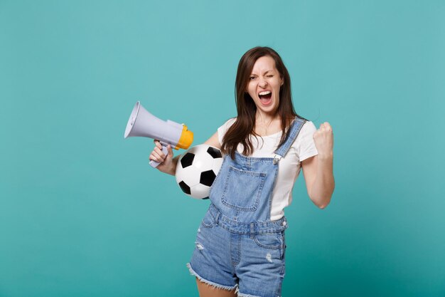 Une jeune femme ravie de supporter l'équipe préférée d'un fan de football avec un mégaphone de ballon de football faisant le geste du vainqueur isolé sur fond bleu turquoise. Émotions des gens, concept de mode de vie de loisirs familiaux sportifs.