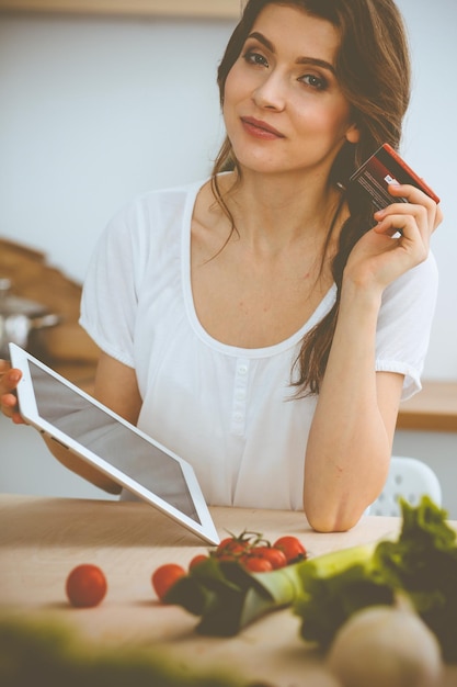 Jeune femme à la recherche d'une nouvelle recette pour cuisiner dans une cuisine. Housewife fait des achats en ligne par tablette et carte de crédit.