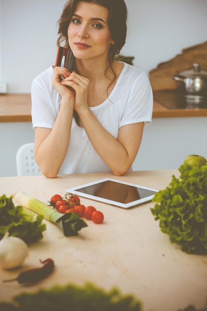 Jeune femme à la recherche d'une nouvelle recette pour cuisiner dans une cuisine. Housewife fait des achats en ligne par tablette et carte de crédit.