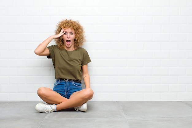 Jeune femme à la recherche de plaisir, étonné et surpris, souriant et réalisant de bonnes et incroyables bonnes nouvelles assis sur un sol