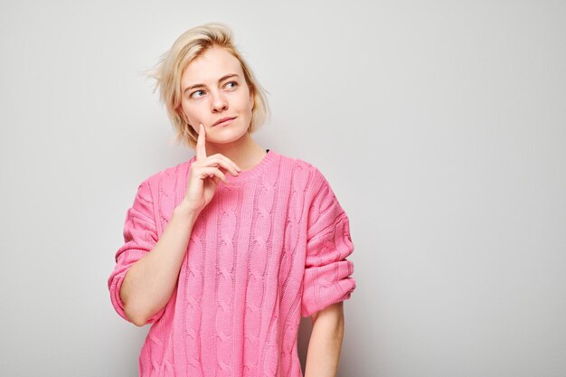 Une jeune femme réfléchie dans un pull rose, la main sur le menton, détournant son regard sur un fond gris.