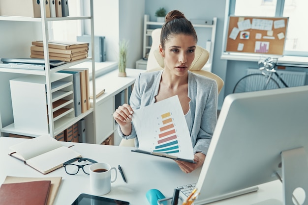 Jeune femme réfléchie dans des vêtements décontractés intelligents travaillant avec des graphiques alors qu'elle était assise au bureau