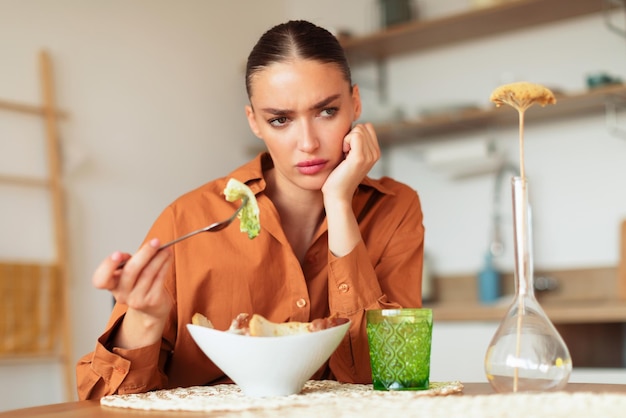 Jeune femme réfléchie mangeant de la salade césar et ayant un manque d'appétit assis à table dans la cuisine