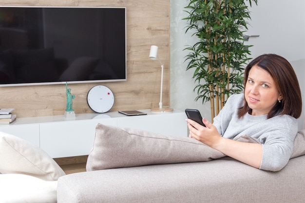 Jeune femme regardant son téléphone intelligent et souriant assis sur le canapé