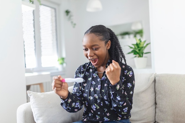 Une jeune femme regardant un test de grossesse dans le bonheur enfin enceinte des femmes noires attrayantes regardant un Test de grossesse et souriant tout en étant assises sur le canapé à la maison