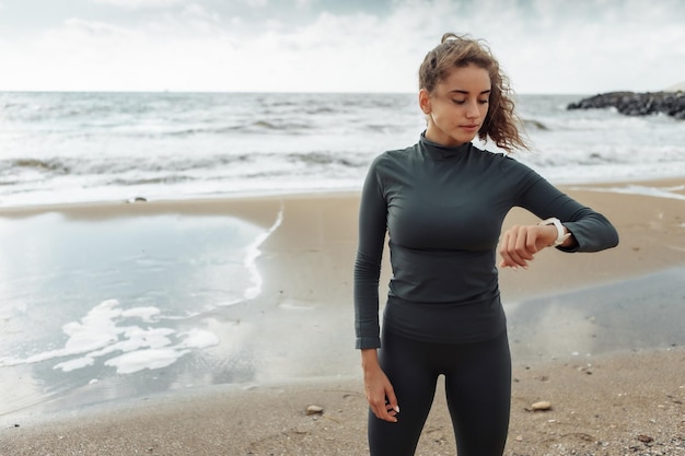 Jeune femme de remise en forme aux cheveux bouclés en tenue de sport regardant la montre sur la plage