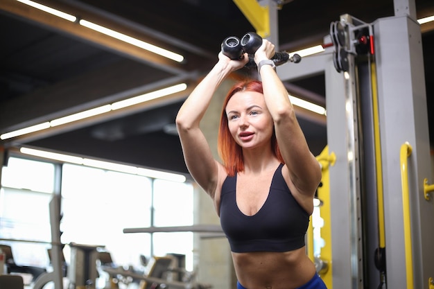 Une jeune femme de remise en forme aux cheveux roux en vêtements de sport entraîne les muscles des triceps dans une machine d'exercice à câble croisé faisant l'extension des bras derrière la tête dans une salle de sport moderne Concept de remise en forme et de musculation