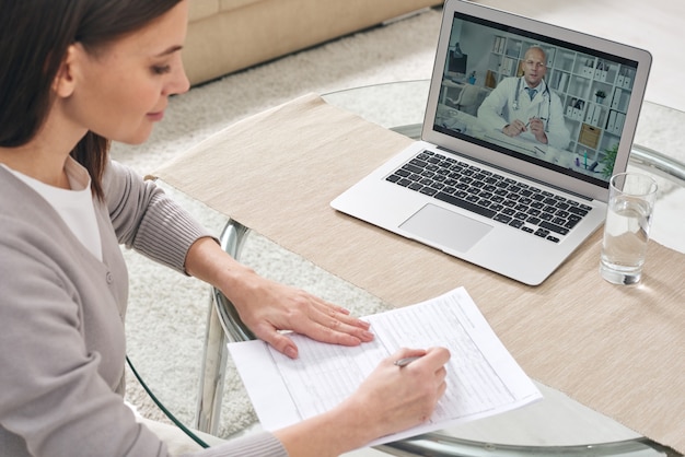 Jeune femme remplissant le formulaire d'assurance maladie et regardant la vidéo en ligne où le médecin donne des conseils médicaux