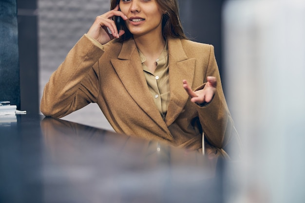 Jeune femme réussie dans le manteau à la mode restant à la réception tout en parlant émotionnellement sur mobile