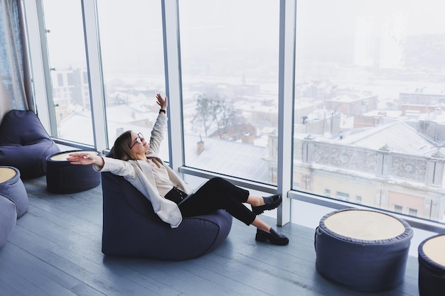 Jeune femme réussie dans une veste souriant et travaillant sur un ordinateur portable dans un bureau moderne avec de grandes fenêtres Repos après le travail Travail à distance