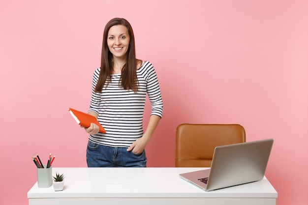 Jeune femme réussie dans des vêtements décontractés tenant un ordinateur portable debout près d'un bureau blanc avec un ordinateur portable contemporain isolé sur fond rose pastel. Concept de carrière d'entreprise de réalisation. Espace de copie.