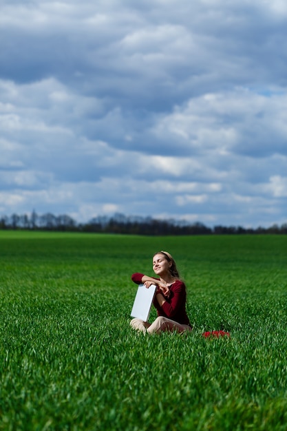 Jeune femme réussie est assise sur l'herbe verte avec un ordinateur portable dans ses mains. Reposez-vous après une bonne journée de travail. Travail sur la nature. Fille étudiante travaillant dans un endroit isolé. Lieu de travail dans la nature