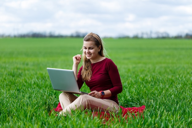 Jeune femme réussie est assise sur l'herbe verte avec un ordinateur portable dans ses mains. Travail sur la nature. Fille étudiante travaillant dans un endroit isolé. De nouvelles idées commerciales