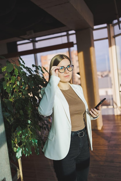 Une jeune femme réussie portant des lunettes et une veste se tient au bureau et parle joyeusement au téléphone Un jeune manager travaille au bureau et gère l'entreprise à distance