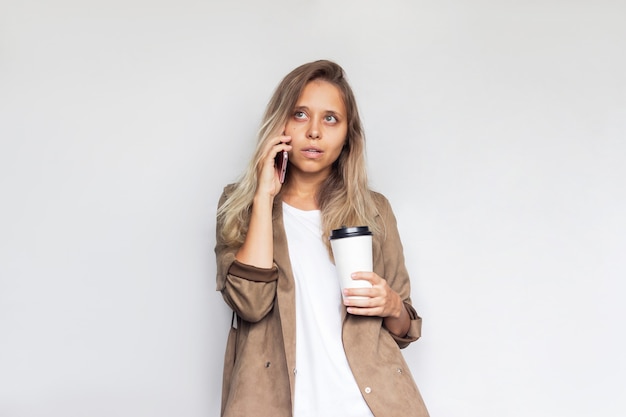 Une jeune femme réussie tient une tasse de café ou de thé en papier blanc parlant sur un téléphone portable