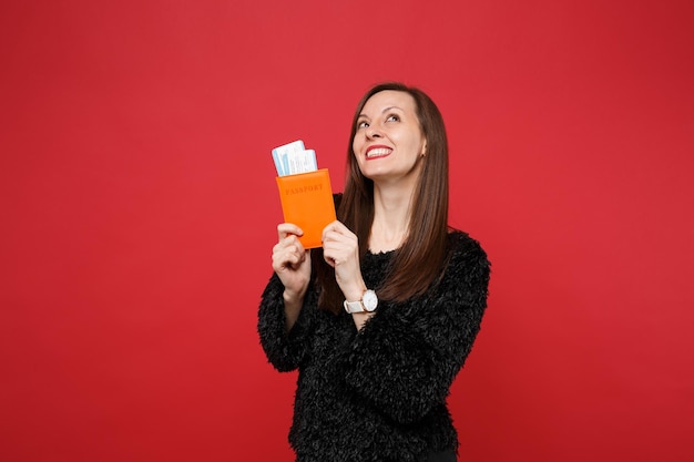 Jeune femme rêveuse en pull de fourrure noire levant les yeux, tenant un passeport, un billet d'embarquement isolé sur fond rouge vif en studio. Les gens émotions sincères, concept de style de vie. Maquette de l'espace de copie.