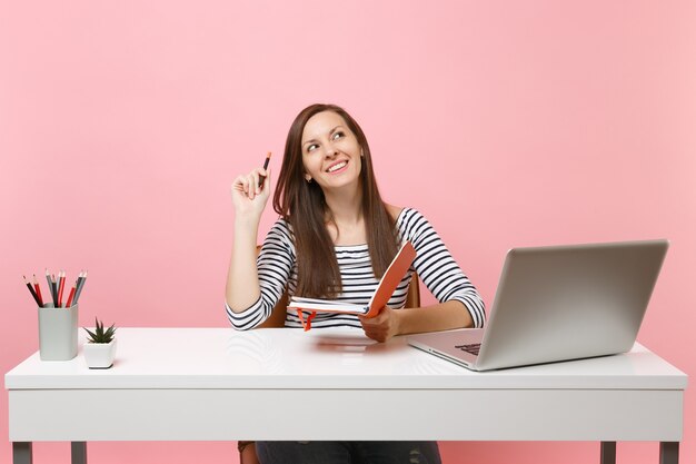 Jeune femme rêveuse recherchant une nouvelle idée en pensant à la recherche d'une nouvelle idée tenant un crayon et un ordinateur portable assis au bureau blanc avec un ordinateur portable pc