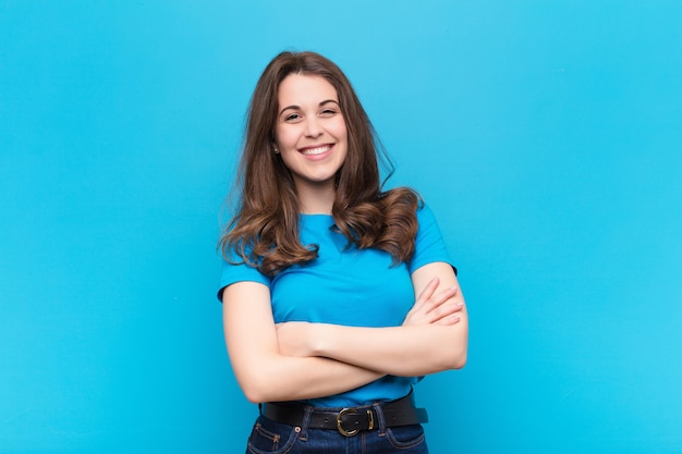 Jeune femme riant joyeusement avec les bras croisés, avec une pose détendue, positive et satisfaite sur le mur bleu