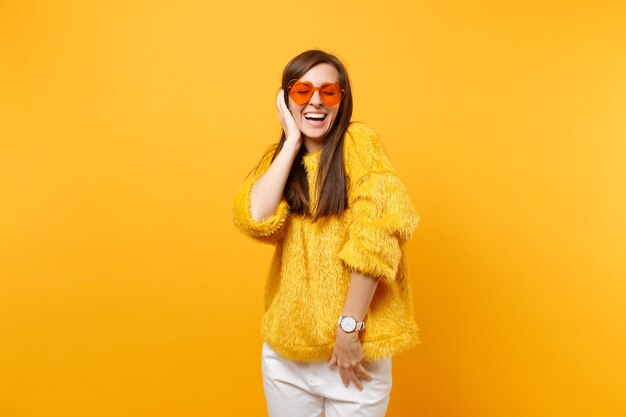 Jeune femme riante aux yeux fermés en pull de fourrure et lunettes orange coeur gardant la main près de la tête isolée sur fond jaune vif. Les gens émotions sincères, concept de style de vie. Espace publicitaire.