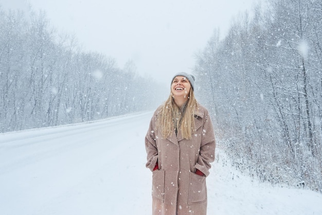 Jeune femme riante dans un manteau et un chapeau dans le parc lors d'une chute de neige en hiver, la blonde se réjouit de la neige