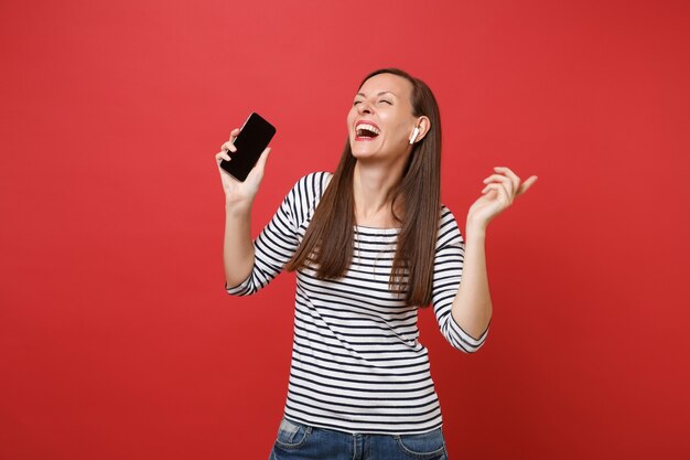 Une jeune femme riante avec des écouteurs sans fil tient un téléphone portable avec un écran vide noir vierge écoutant de la musique isolée sur fond rouge. Les gens émotions sincères, concept de style de vie. Maquette de l'espace de copie.