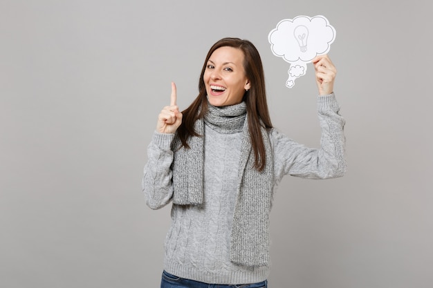 Jeune femme riante en pull, écharpe tenant l'index avec une nouvelle idée géniale, disons nuage avec ampoule isolé sur fond gris. Concept de mode de vie sain des émotions des gens de la saison froide.