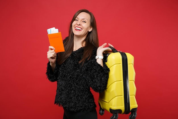 Jeune femme riante en pull de fourrure noire tenant une valise, un passeport, un billet d'embarquement isolé sur fond rouge vif en studio. Les gens émotions sincères, concept de style de vie. Maquette de l'espace de copie.