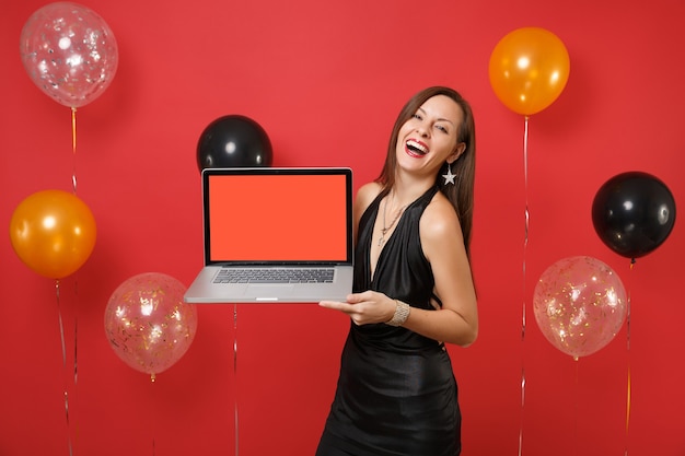 Jeune Femme Riante En Robe Noire Célébrant, Tenant Un Ordinateur Portable Avec Un écran Vide Noir Vide Sur Des Ballons à Air De Fond Rouge Vif. Bonne Année, Concept De Fête De Vacances De Maquette D'anniversaire.