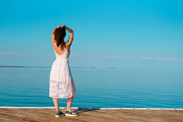 Une jeune femme en robe blanche se dresse sur une jetée en bois avec ses mains sur sa tête