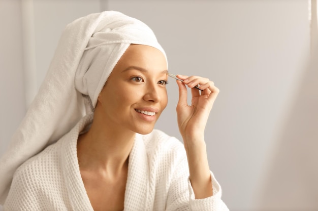 Jeune femme en robe blanche et avec une serviette sur la tête s'épile les sourcils avec des pincettes Nettoyage et soins du corps sains après la douche