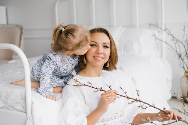 Jeune femme en robe blanche tient une branche de Pâques dans ses mains et s'assied à la maison près du lit sa fille l'embrasse sur la joue