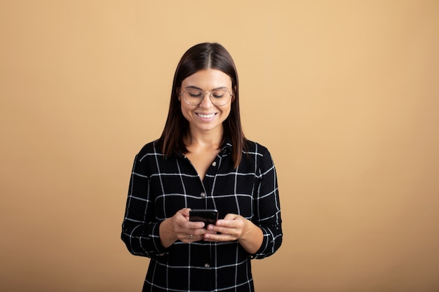 Une jeune femme en robe à carreaux se dresse sur un fond orange et joue avec son téléphone