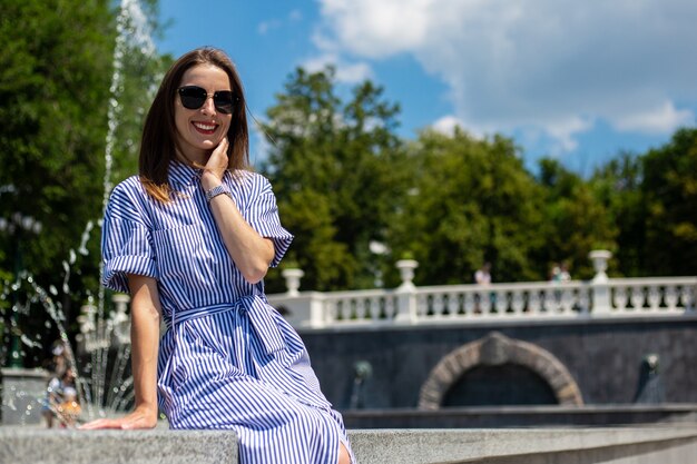 Jeune femme en robe et lunettes se trouve près d'une fontaine dans le parc.