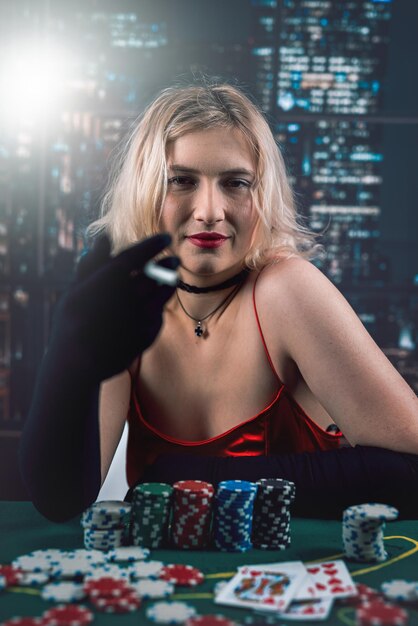 Jeune femme en robe rouge fume une cigarette souffle de la fumée et joue aux cartes au poker de table de poker